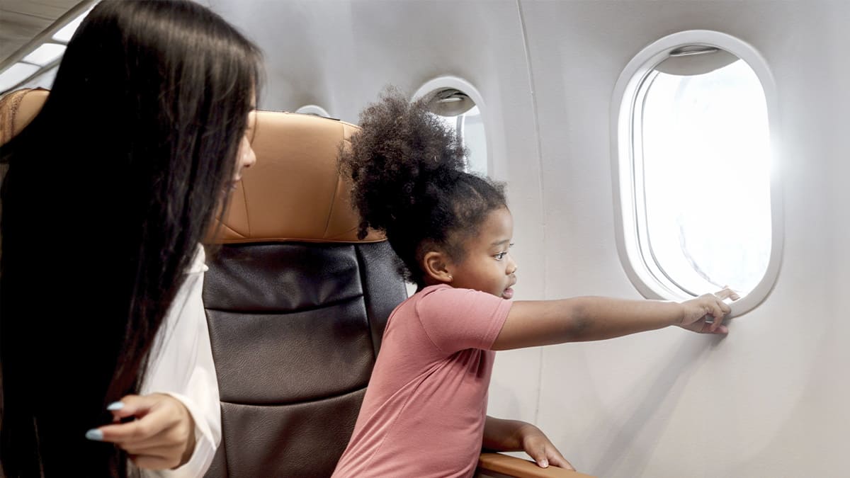 Menina curiosa apontando para fora da janela de um avião enquanto uma mulher a observa. (Créditos: Depositphotos)