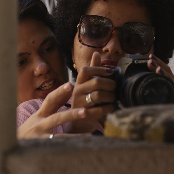 Instrutora orientando mulher com baixa visão no manuseio de câmera fotográfica, durante oficina de fotografia Um Outro Olhar. (Créditos: Alberto Contarato & Cia Poéticas da Cena Contemporânea)