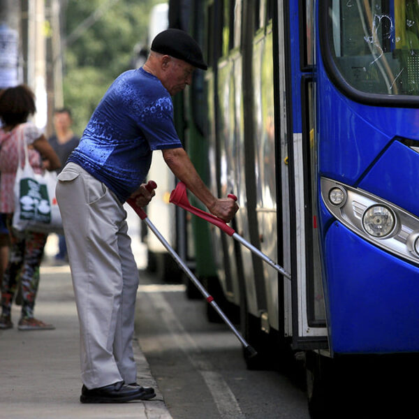 Homem idoso com muletas embarcando em um ônibus azul de número 10134, enquanto outras pessoas aguardam na calçada. Cena retrata mobilidade urbana. (Foto: Depositphotos)