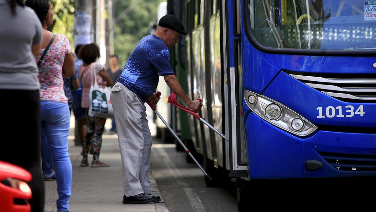 Homem idoso com muletas embarcando em um ônibus azul de número 10134, enquanto outras pessoas aguardam na calçada. Cena retrata mobilidade urbana.(Foto: Depositphotos)