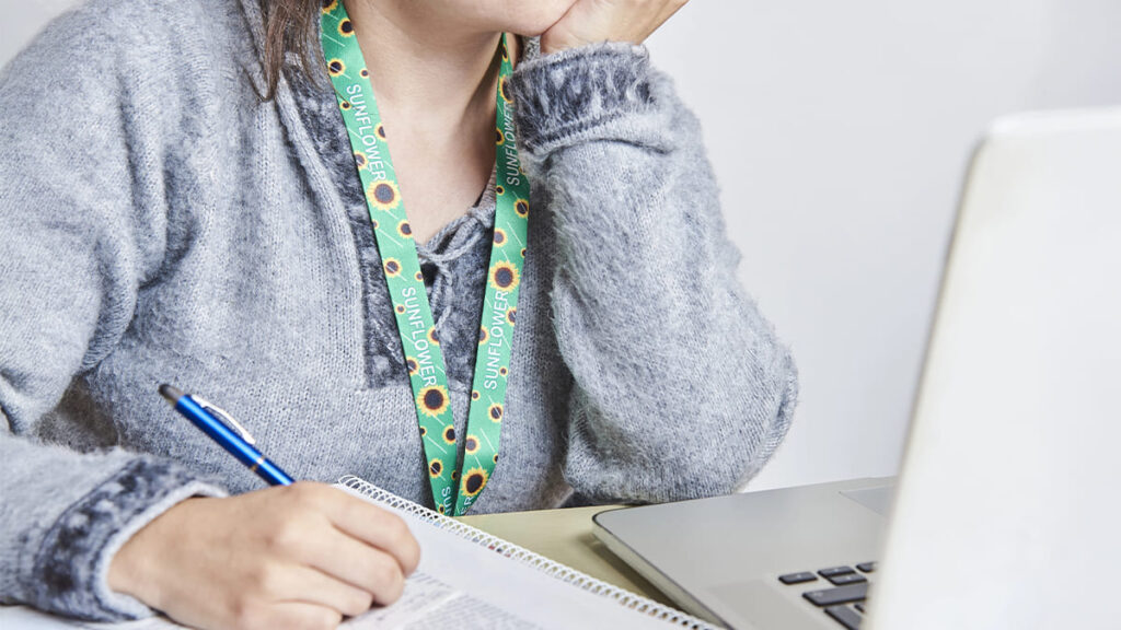 Pessoa usando cordão girassol, símbolo de deficiência oculta, como no caso de estudantes com autismo, enquanto escreve em um caderno diante de um notebook. (Créditos: Depositphotos)