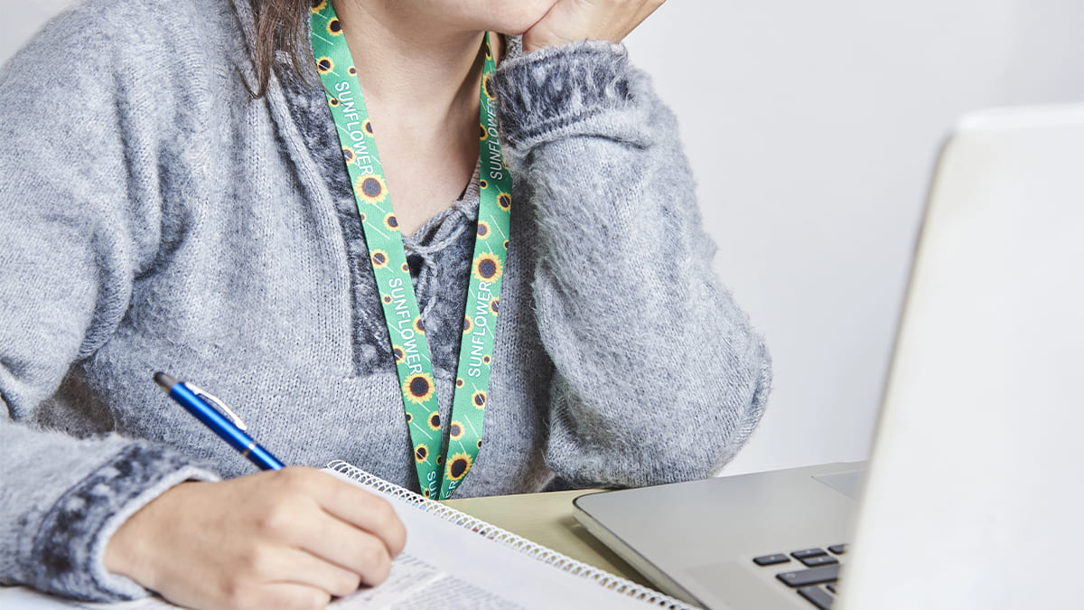 Pessoa usando cordão girassol, símbolo de deficiência oculta, como no caso de estudantes com autismo, enquanto escreve em um caderno diante de um notebook. (Créditos: Depositphotos)