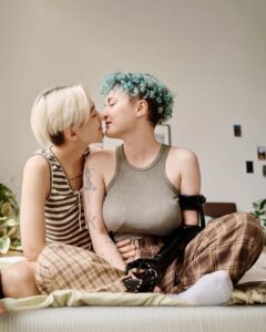 Duas mulheres sentadas na cama se beijam com afeto, uma delas usando prótese no braço direito, mostrando que amor não vê deficiência.  
