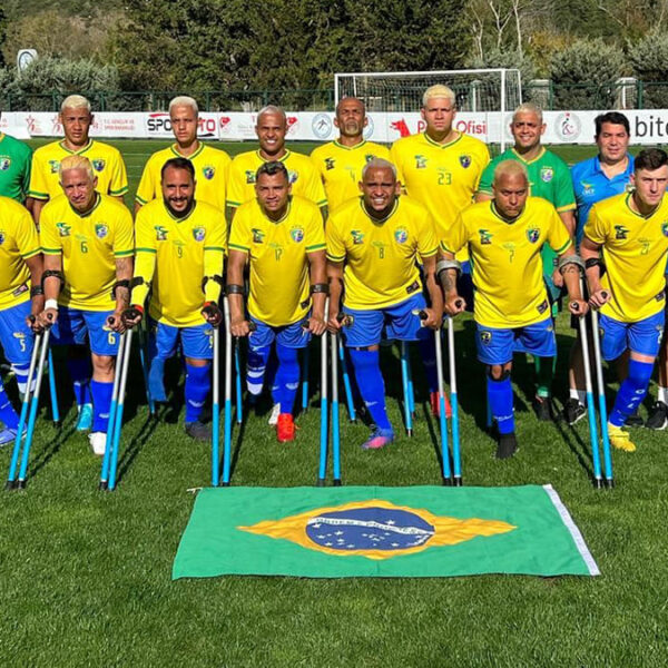Seleção brasileira de futebol de amputados posa em campo com uniforme amarelo e azul, ao lado da comissão técnica, atrás de uma bandeira do Brasil.
