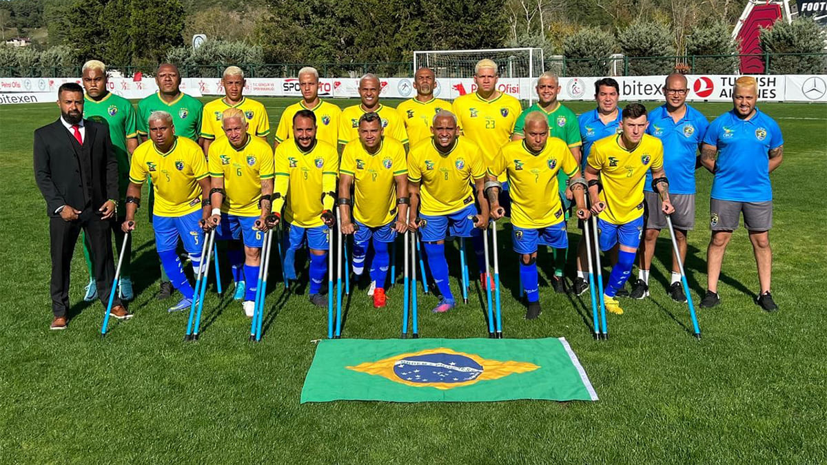 Seleção brasileira de futebol de amputados posa em campo com uniforme amarelo e azul, ao lado da comissão técnica, atrás de uma bandeira do Brasil.