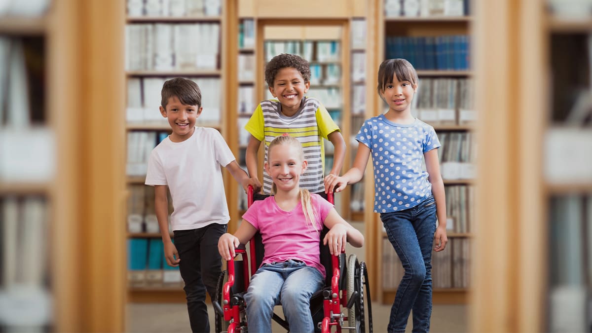 Crianças sorridentes em uma biblioteca, incluindo uma menina em cadeira de rodas cercada por colegas, representando inclusão e o direito de ser amigo. (Créditos: Depositphotos)