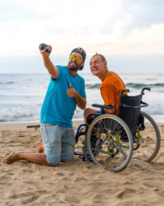 Homem em cadeira de rodas e amigo tirando selfie na praia, com o mar ao fundo, representando o direito de ser amigo e lazer com acessibilidade.