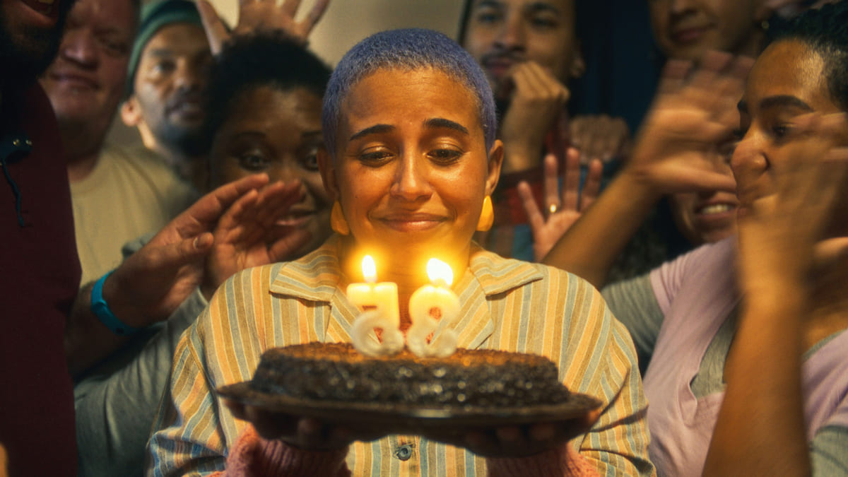 Pessoa segura bolo com velas de 35 anos, rodeada por amigos. Cena do filme "Nem Toda História de Amor Acaba em Morte", protagonizado pela atriz surda Gabriela Grigolom. (Créditos: Beija Flor Filmes)
