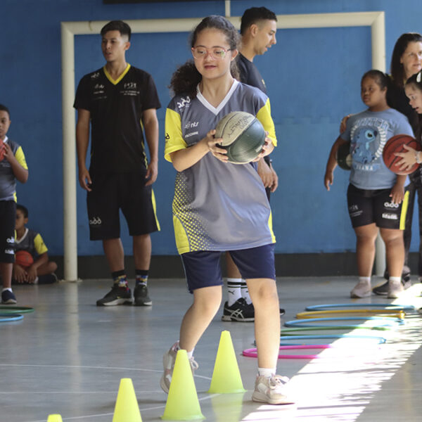 Menina com deficiência participa de atividade esportiva em quadra, segurando bola e caminhando entre cones. Ao fundo, colegas e instrutores do projeto da ADD. (Créditos: Divulgação)