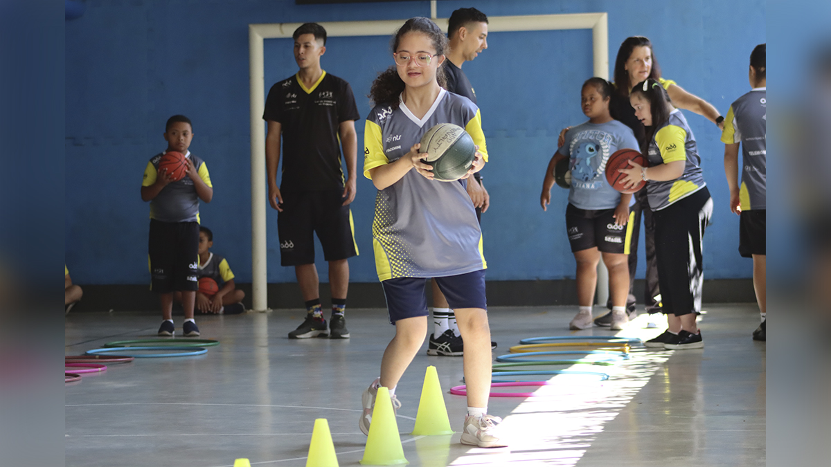 Menina com deficiência participa de atividade esportiva em quadra, segurando bola e caminhando entre cones. Ao fundo, colegas e instrutores do projeto da ADD. (Créditos: Divulgação)