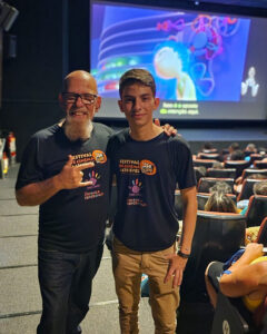 Dois homens com camisetas do Festival de Cinema Acessível Kids posam juntos em sala de cinema durante exibição de “Divertida Mente” com legenda e intérprete de Libras.
