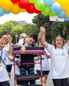Jovem em andador é apoiado por participantes sob arco de balões coloridos na Caminhada Inclusiva Mover-se 2024 em São Paulo.