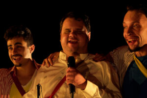 Homem com microfone canta sorridente ao lado de dois colegas em apresentação musical no palco – Cia Poéticas, realizadora da oficina de teatro para homens cegos.