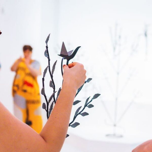 Uma mulher de óculos, vista de perfil, interage com escultura de metal semelhante a um galho com um pássaro de origami no topo, em uma galeria de arte com fundo branco. (Foto: Lusaneyes)