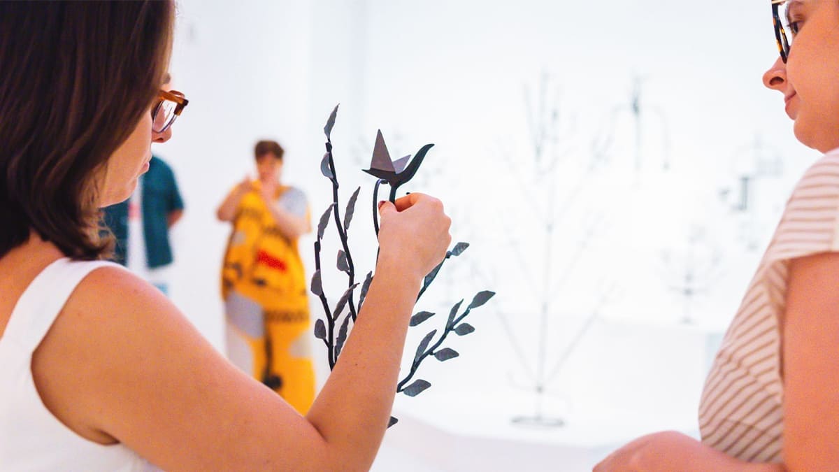 Uma mulher de óculos, vista de perfil, interage com escultura de metal semelhante a um galho com um pássaro de origami no topo, em uma galeria de arte com fundo branco. (Foto: Lusaneyes)