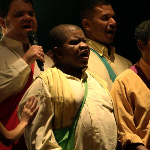 Grupo de homens cegos canta no palco, com microfone em mãos e iluminação dramática durante apresentação musical. (Créditos: Divulgação)