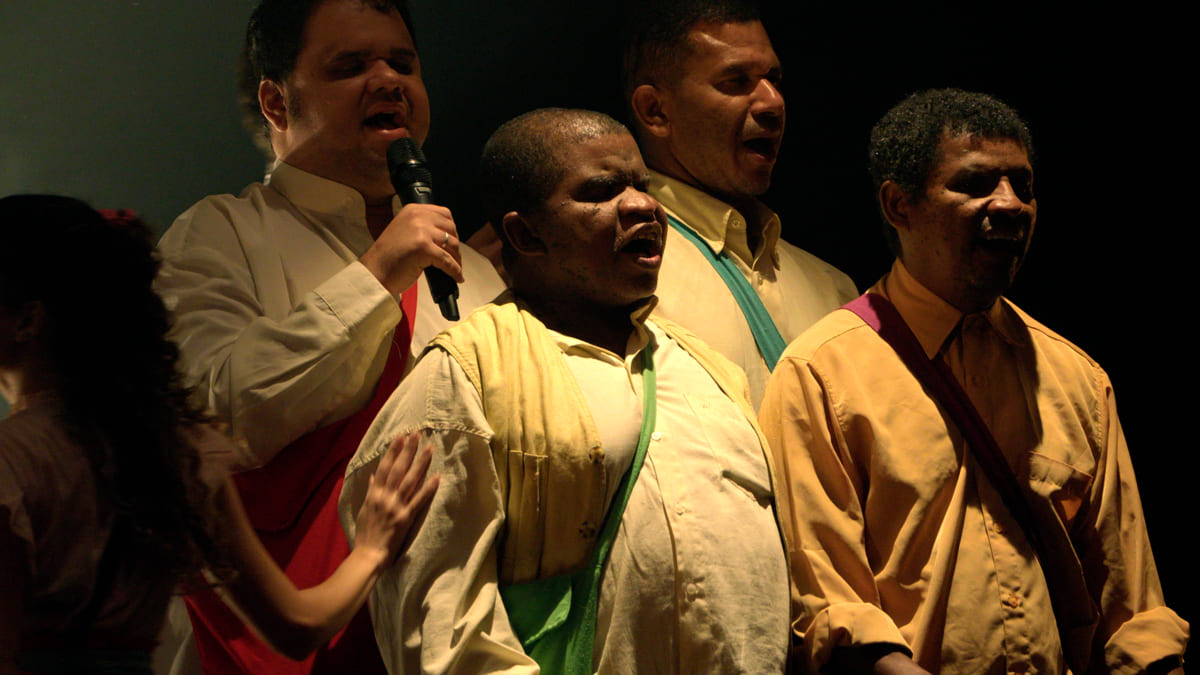Grupo de homens cegos canta no palco, com microfone em mãos e iluminação dramática durante apresentação musical. (Créditos: Divulgação)