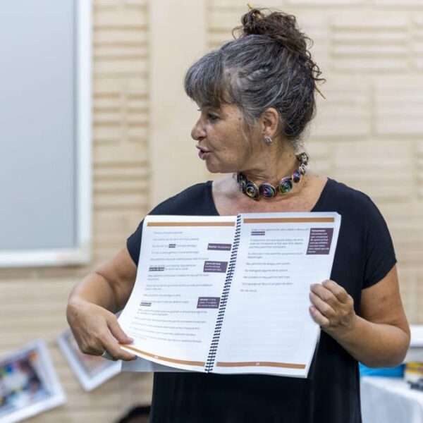 Carla Mauch, coordenadora da ONG Mais Diferenças, segura apostila aberta e explica conteúdo durante atividade formativa. (Créditos: Divulgação / Mais Diferenças)