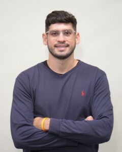 Ronaldo Tenório, especialista em acessibilidade, posa para a foto. Ele é um homem com cabelo escuro e barba, usa óculos e uma camisa azul.