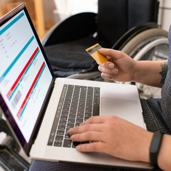 Pessoa usuária de cadeira de rodas realiza uma compra online, segurando um cartão de crédito amarelo enquanto digita em um laptop que exibe uma página de transações. (Foto: Depositphotos)