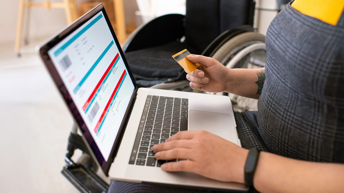Pessoa usuária de cadeira de rodas realiza uma compra online, segurando um cartão de crédito amarelo enquanto digita em um laptop que exibe uma página de transações. (Foto: Depositphotos)
