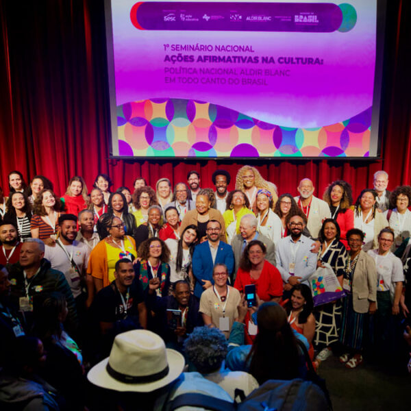 Foto oficial com um grupo diverso de participantes do 1º Seminário Nacional de Ações Afirmativas na Cultura, em frente a um telão. (Foto: Filipe Araújo/MinC)