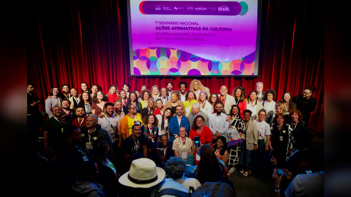 Foto oficial com um grupo diverso de participantes do 1º Seminário Nacional de Ações Afirmativas na Cultura, em frente a um telão. (Foto: Filipe Araújo/MinC)