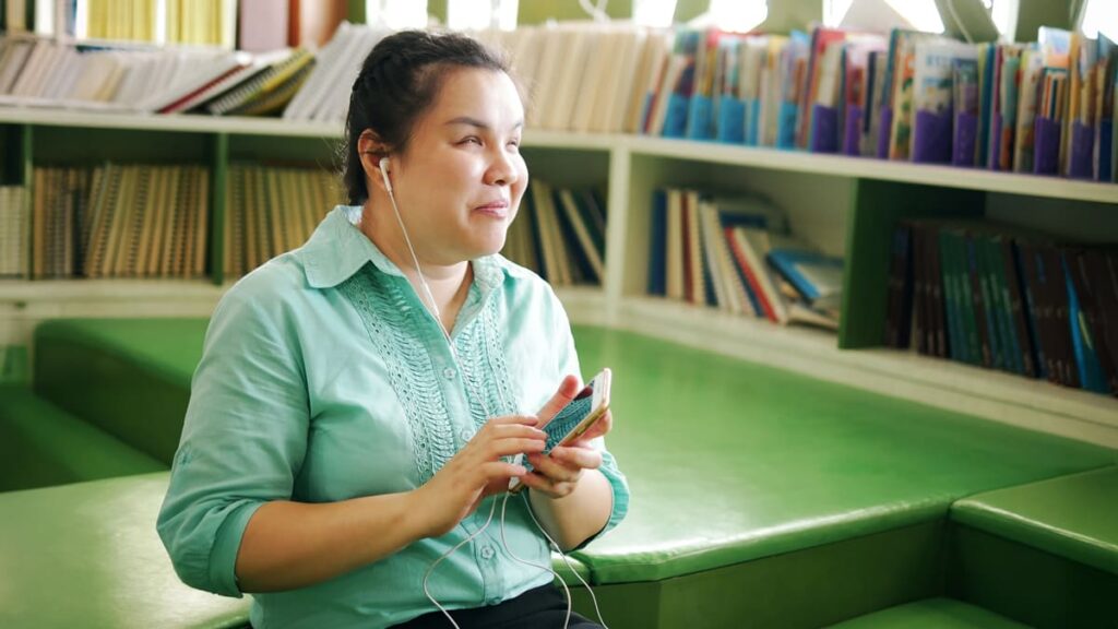 Mulher com deficiência visual sorri ao ouvir um audiobook em seu celular, sentada na área de leitura de uma biblioteca moderna. Foto: Depositphotos