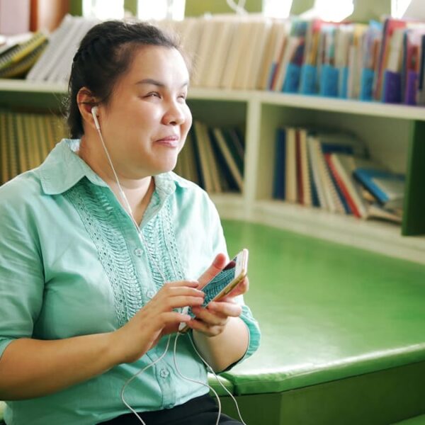 Mulher com deficiência visual sorri ao ouvir um audiobook em seu celular, sentada na área de leitura de uma biblioteca com estante de livros ao fundo. (Foto: Depositphotos)