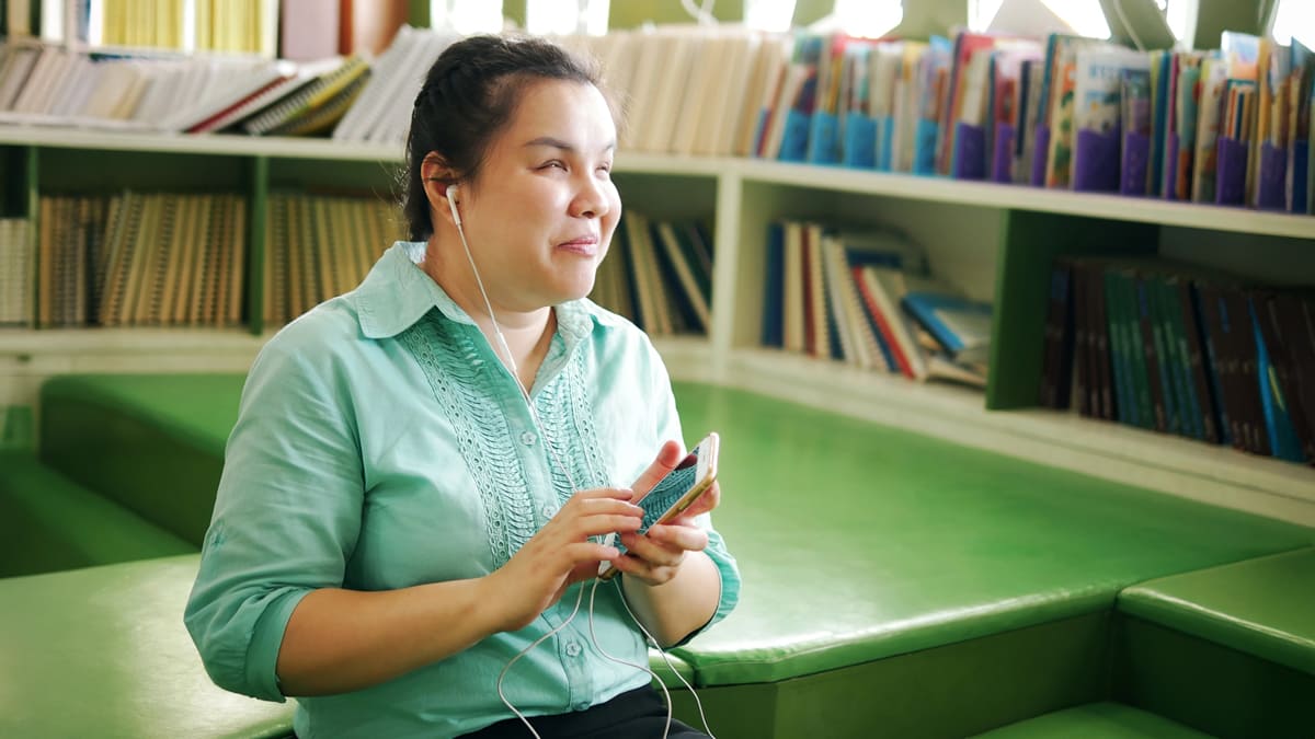 Mulher com deficiência visual sorri ao ouvir um audiobook em seu celular, sentada na área de leitura de uma biblioteca moderna. Foto: Depositphotos