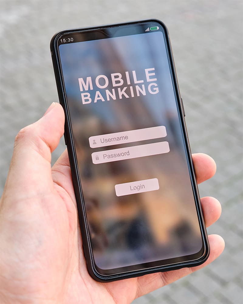 Mão segura smartphone exibindo a tela de login de um aplicativo de "Mobile Banking", com campos para usuário e senha visíveis, e a falta de acessibilidade em apps bancários.
