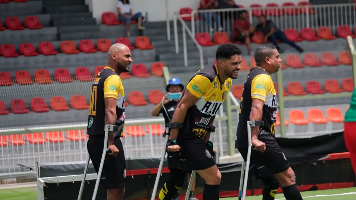 Três jogadores de futebol para amputados, vestindo uniformes preto e amarelo, caminham no gramado de um estádio usando muletas, com a arquibancada vermelha ao fundo. (Foto: Acervo/Acre Clube)