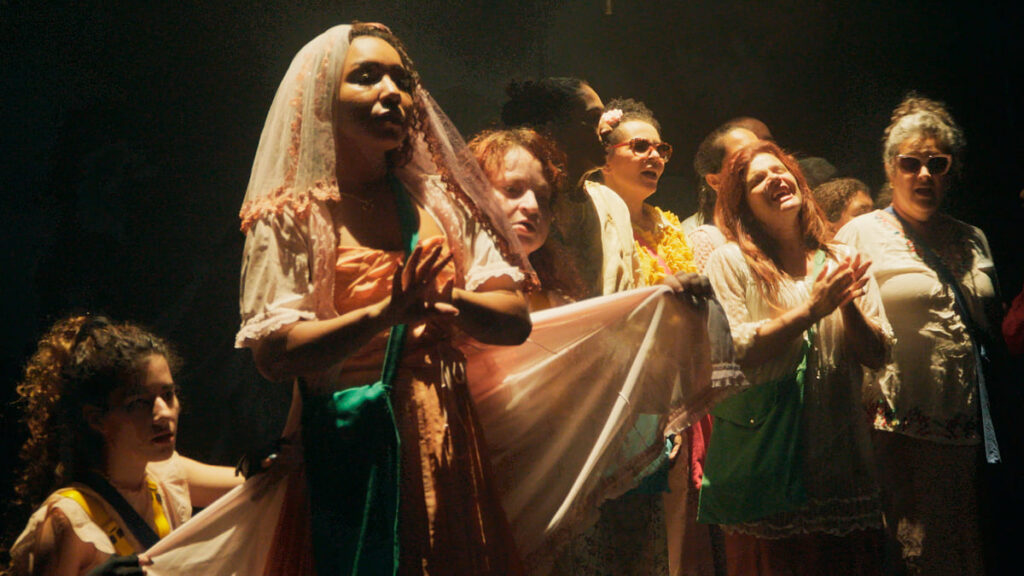Close-up de um grupo de cantores cegos e atores no palco, com uma mulher em primeiro plano usando um véu, todos cantando. (Foto: Julio Gabriel/Cia Poéticas da Cena Contemporânea)