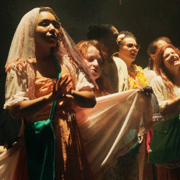 Close-up de um grupo de cantores cegos e atores no palco, com uma mulher em primeiro plano usando um véu, todos cantando. (Foto: Julio Gabriel/Cia Poéticas da Cena Contemporânea)