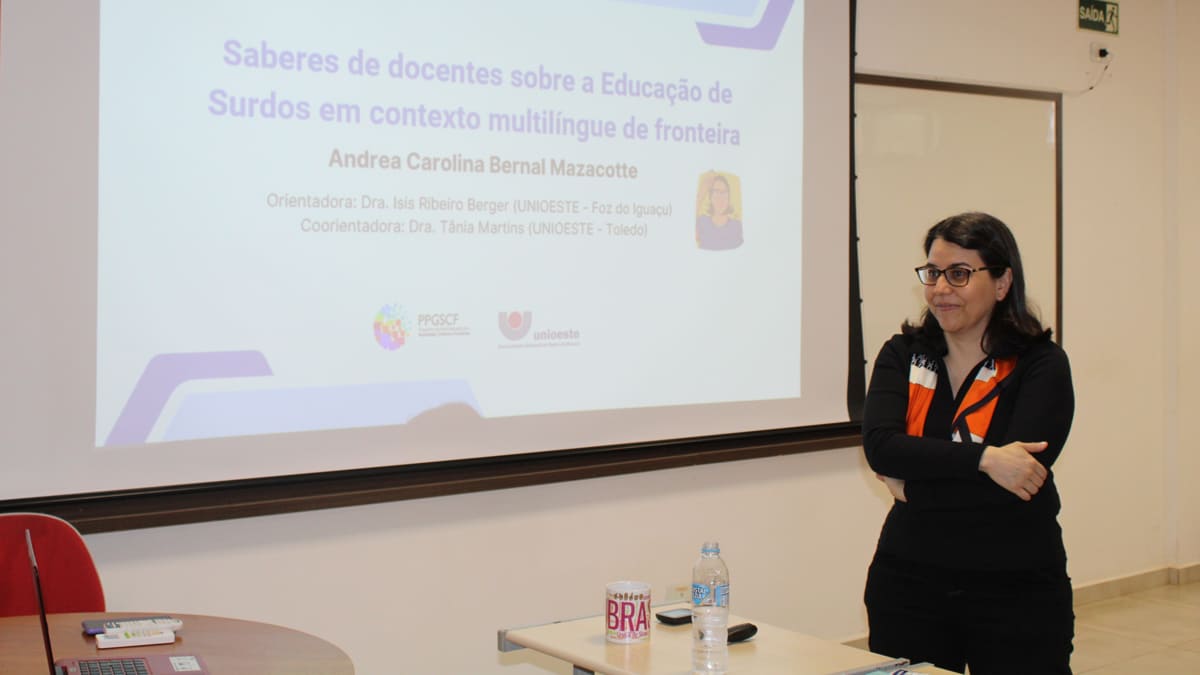 Professora Andréa Mazacotte, uma mulher surda de óculos e cabelos escuros, gesticula em Libras durante a apresentação de sua tese de doutorado na Unioeste. (Foto: Divulgação)