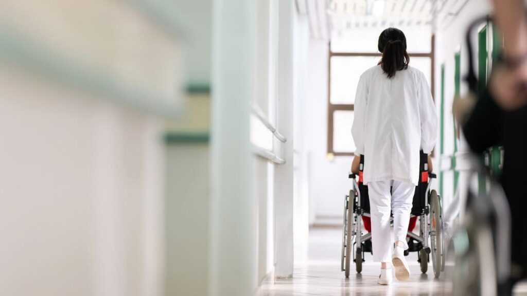 Vista de costas de uma profissional de saúde empurrando um paciente em uma cadeira de rodas por um corredor de hospital iluminado. (Foto: Depositphotos)