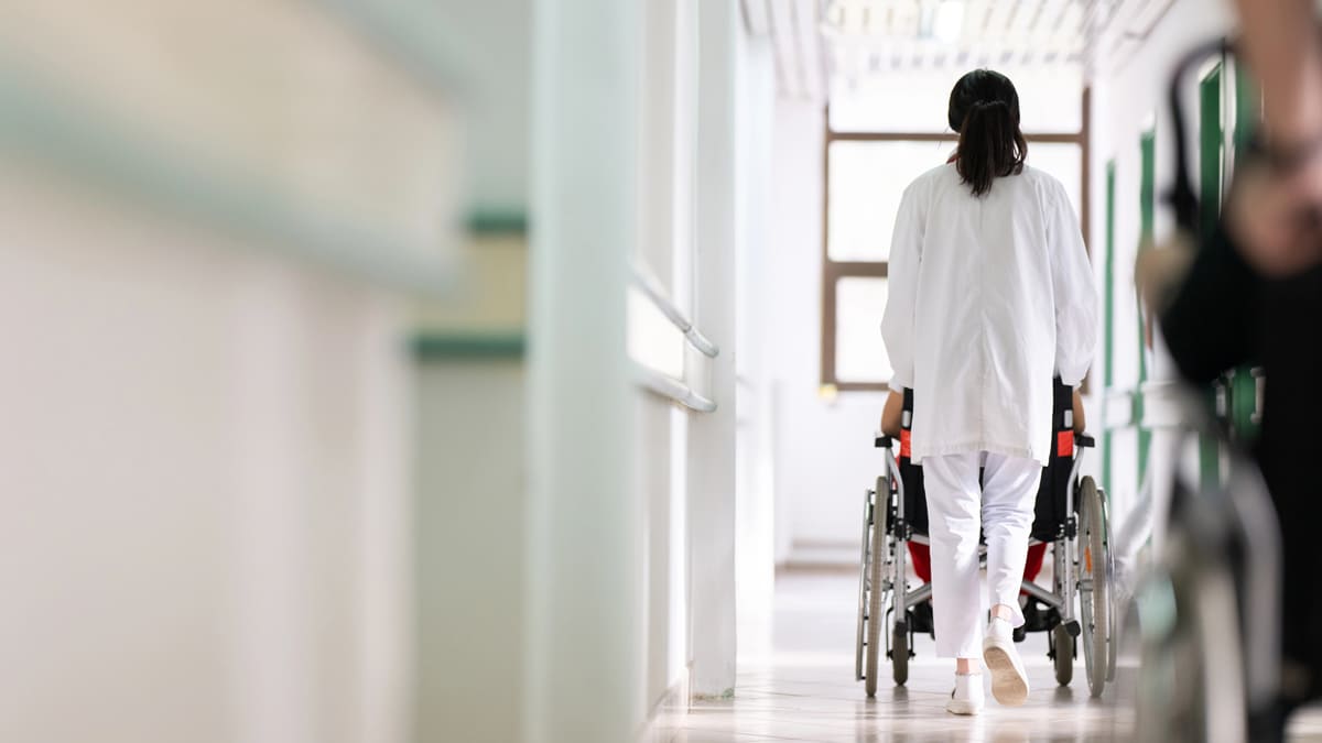 Vista de costas de uma profissional de saúde empurrando um paciente em uma cadeira de rodas por um corredor de hospital iluminado. (Foto: Depositphotos)