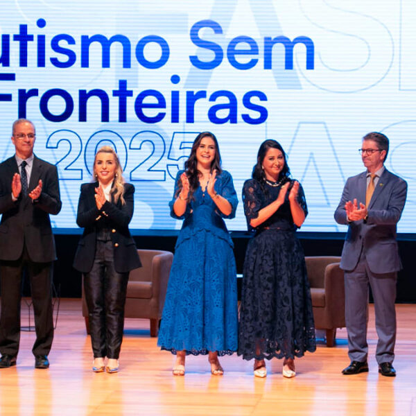 Sete palestrantes em um palco aplaudem em frente a um telão com o título do evento "Autismo Sem Fronteiras 2025". (Foto: Reprodução)