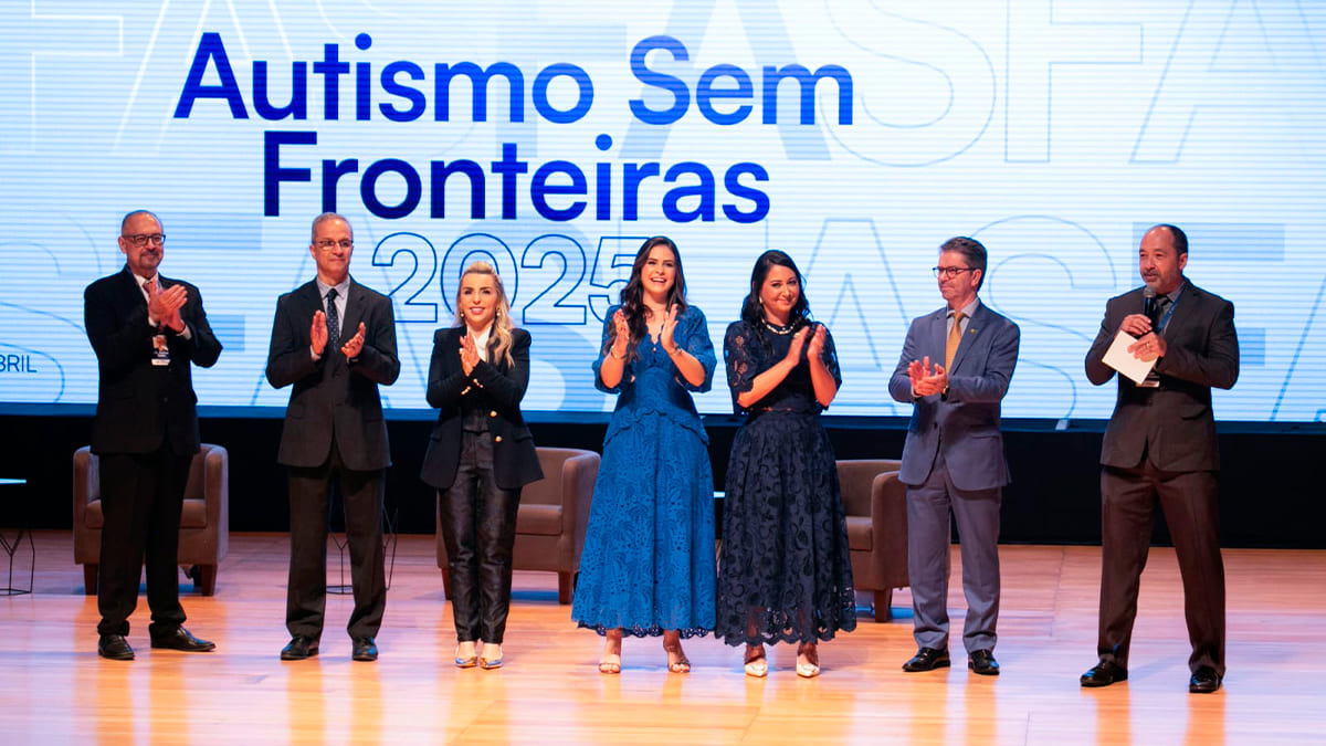 Sete palestrantes em um palco aplaudem em frente a um telão com o título do evento "Autismo Sem Fronteiras 2025". (Foto: Reprodução)