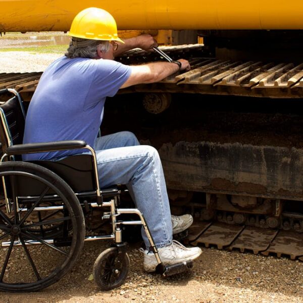 Trabalhador com capacete, sentado em uma cadeira de rodas, realiza a manutenção na esteira de um trator amarelo. (Créditos: Shutterstock)