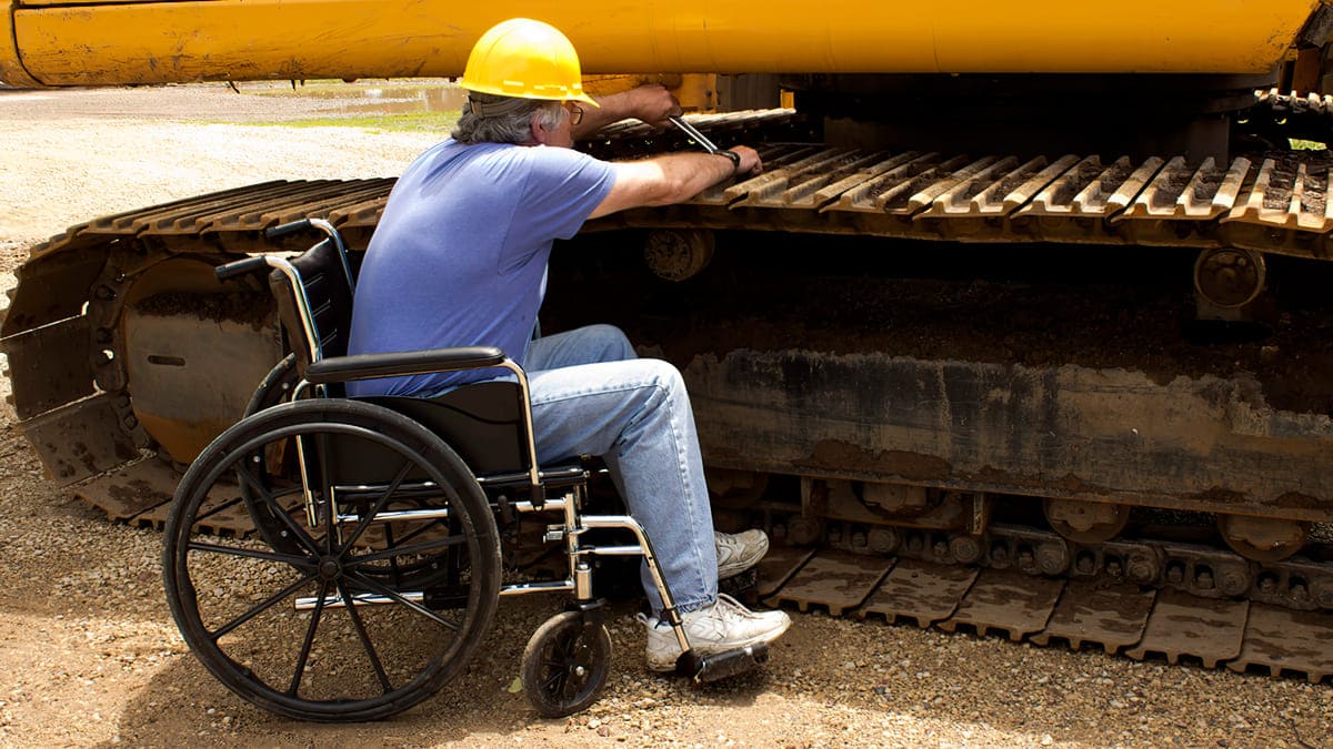 Trabalhador com capacete, sentado em uma cadeira de rodas, realiza a manutenção na esteira de um trator amarelo. (Créditos: Shutterstock)