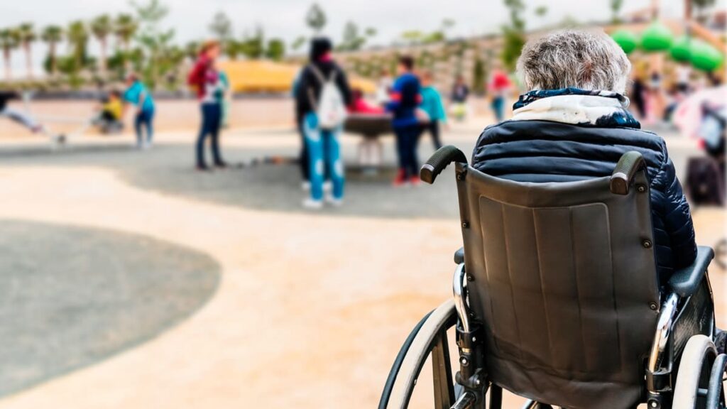 Pessoa idosa em cadeira de rodas, de costas, observa um grupo de pessoas socializando em um parque público. (Foto: Depositphotos)