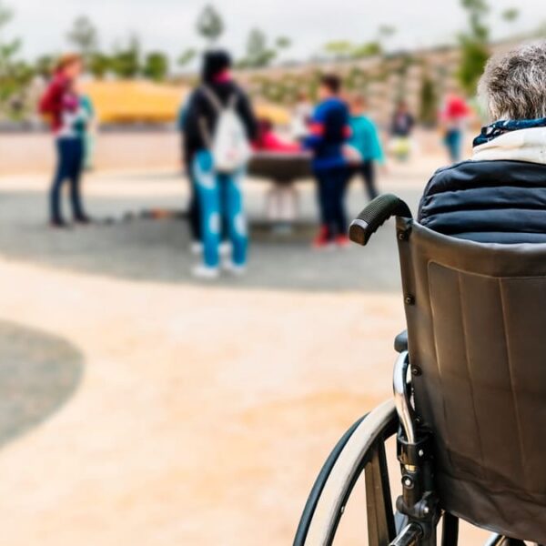 Pessoa idosa em cadeira de rodas, de costas, observa um grupo de pessoas socializando em um parque público. (Foto: Depositphotos)