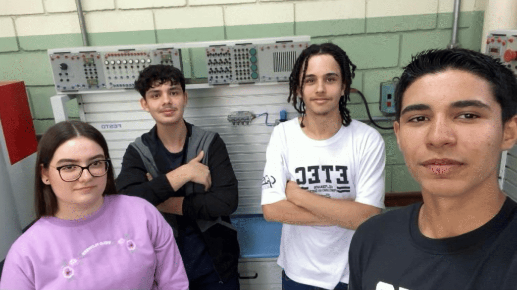 Quatro estudantes, uma mulher e três homens, no laboratório de tecnologia com painel eletrônico ao fundo. (Foto: Divulgação)