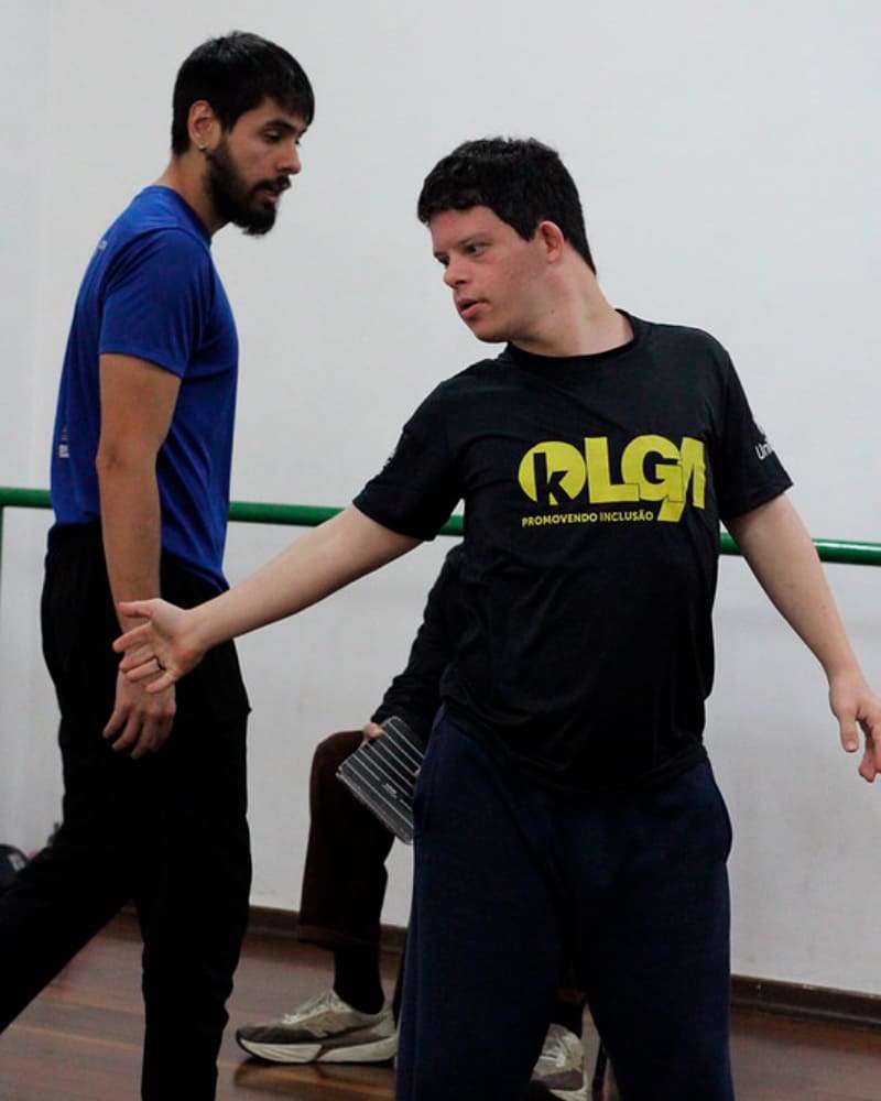 Jovem com síndrome de Down, vestindo uma camiseta preta, dança com um professor em uma sala de ensaio com piso de madeira.