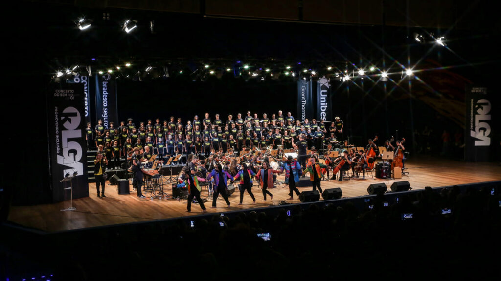 Visão ampla de um palco, uma orquestra, o coral e dançarinos do Instituto Olga Kos durante a apresentação do Concerto do Bem. (Foto: Divulgação/IOK)