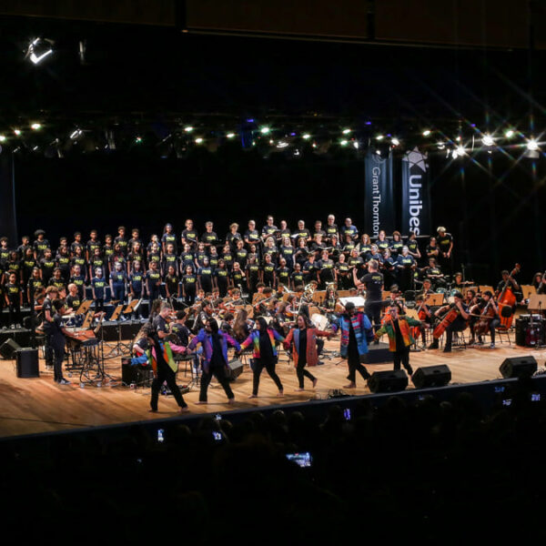 Visão ampla de um palco, uma orquestra, o coral e dançarinos do Instituto Olga Kos durante a apresentação do Concerto do Bem. (Foto: Divulgação/IOK)