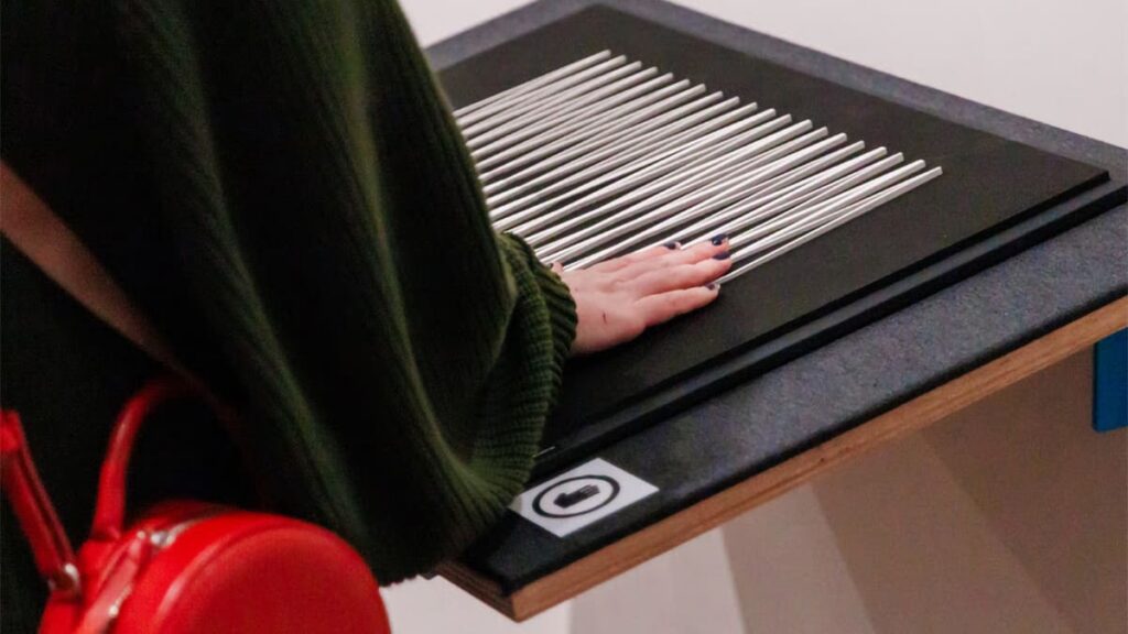 Mão de uma pessoa com unhas escuras toca uma obra de arte tátil, composta por várias hastes de metal paralelas sobre uma base preta. (Foto: @Lusaneyes)