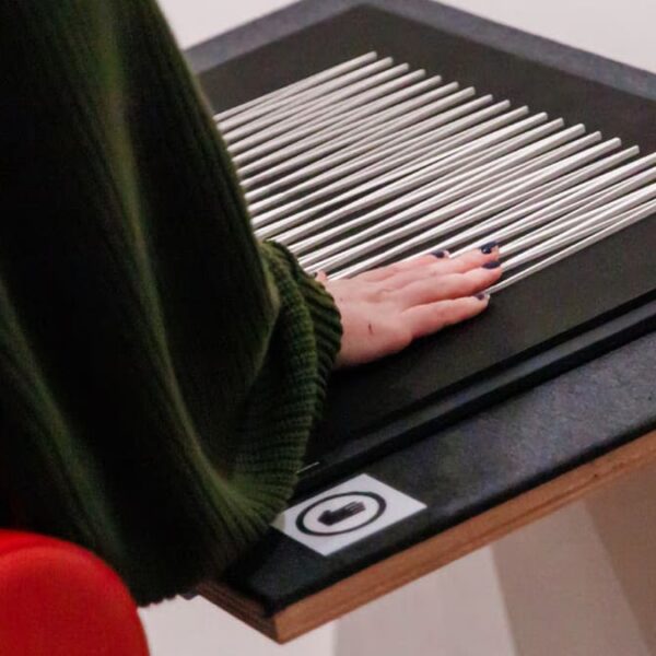 Mão de uma pessoa com unhas escuras toca uma obra de arte tátil, composta por várias hastes de metal paralelas sobre uma base preta. (Foto: @Lusaneyes)