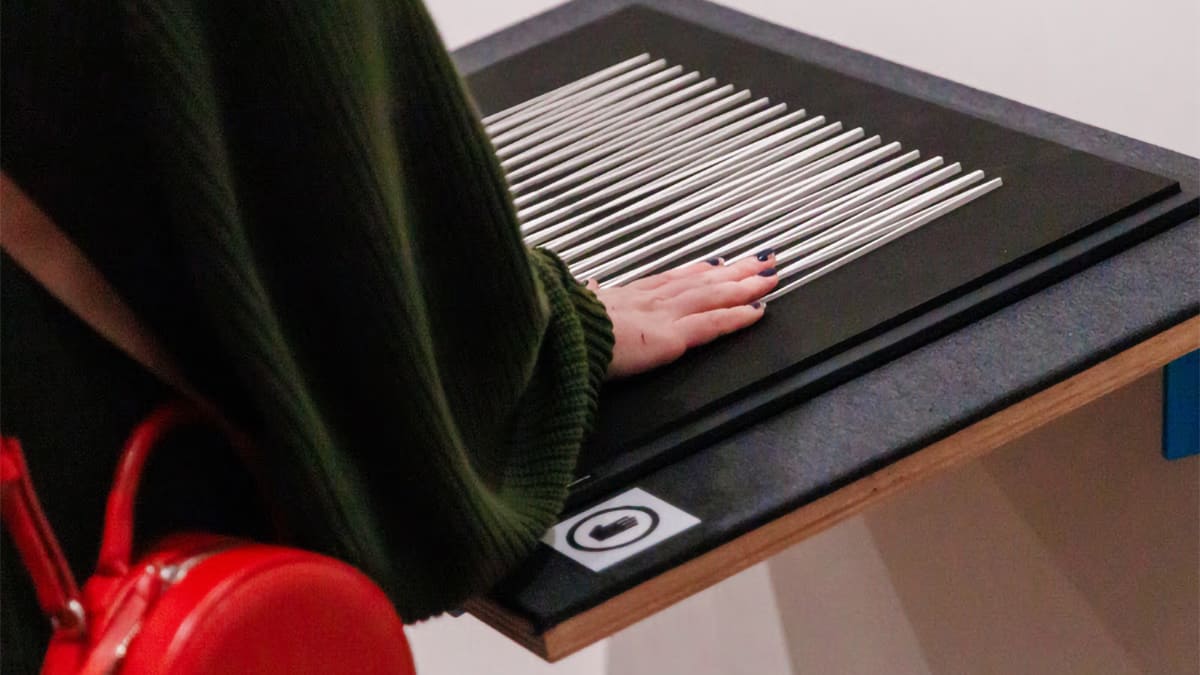 Mão de uma pessoa com unhas escuras toca uma obra de arte tátil, composta por várias hastes de metal paralelas sobre uma base preta. (Foto: @Lusaneyes)